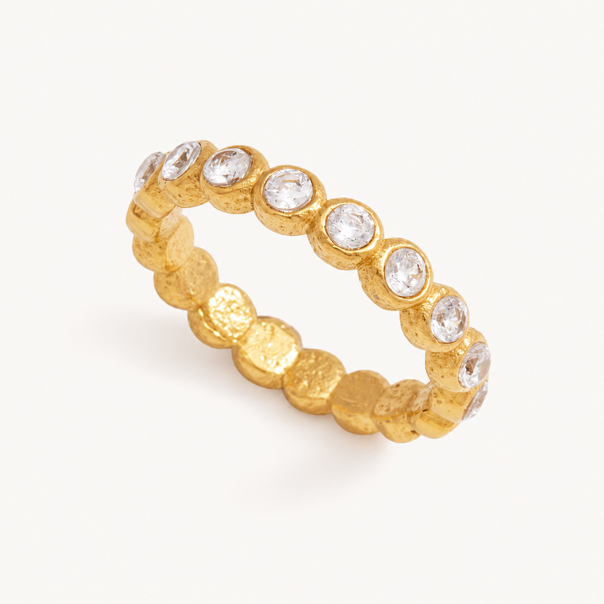 Lucetta gold textured eternity ring with clear bezel-set zircon stones on a warm ivory background.